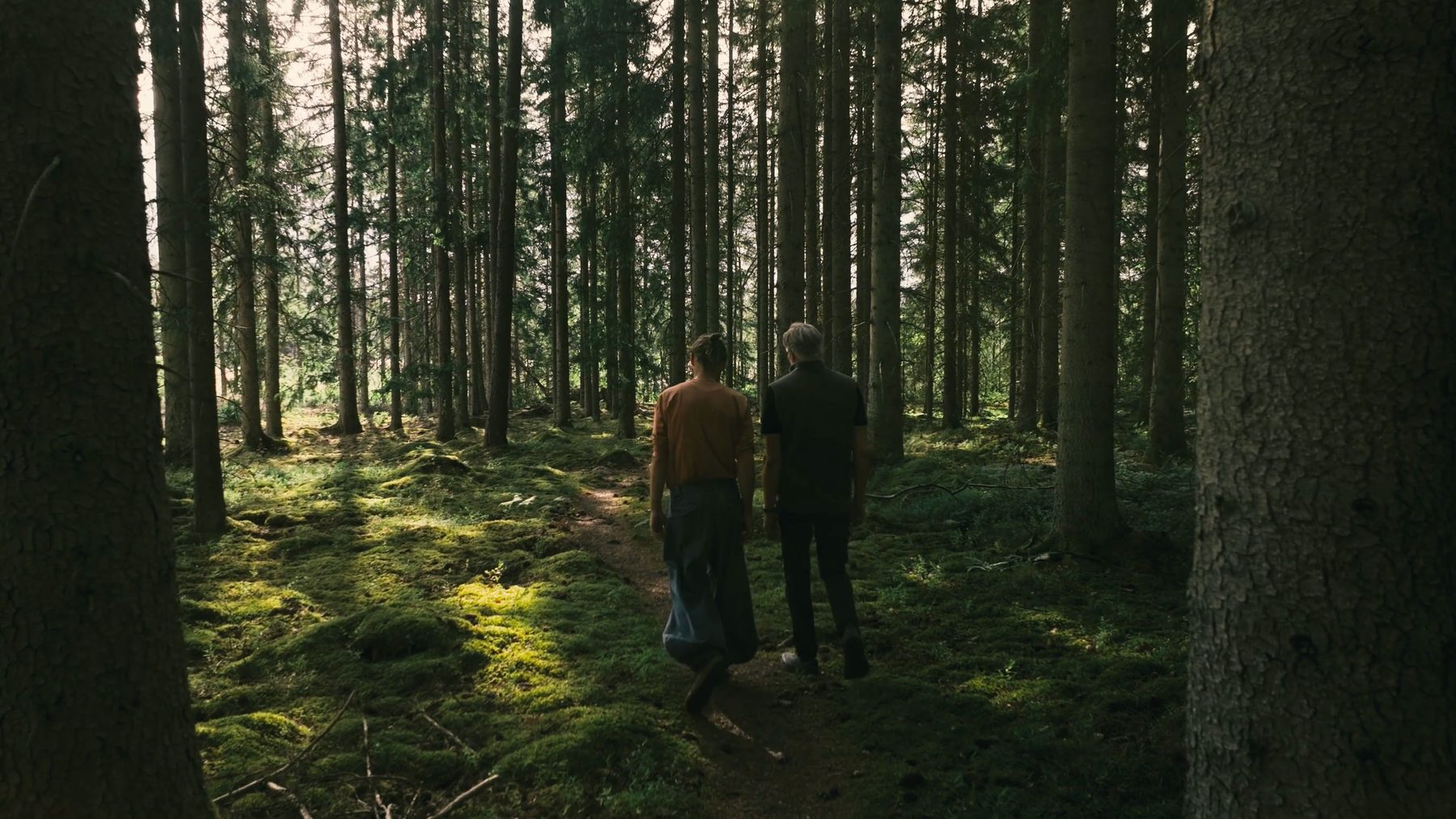Två personer går på en stig genom en mossig granskog
